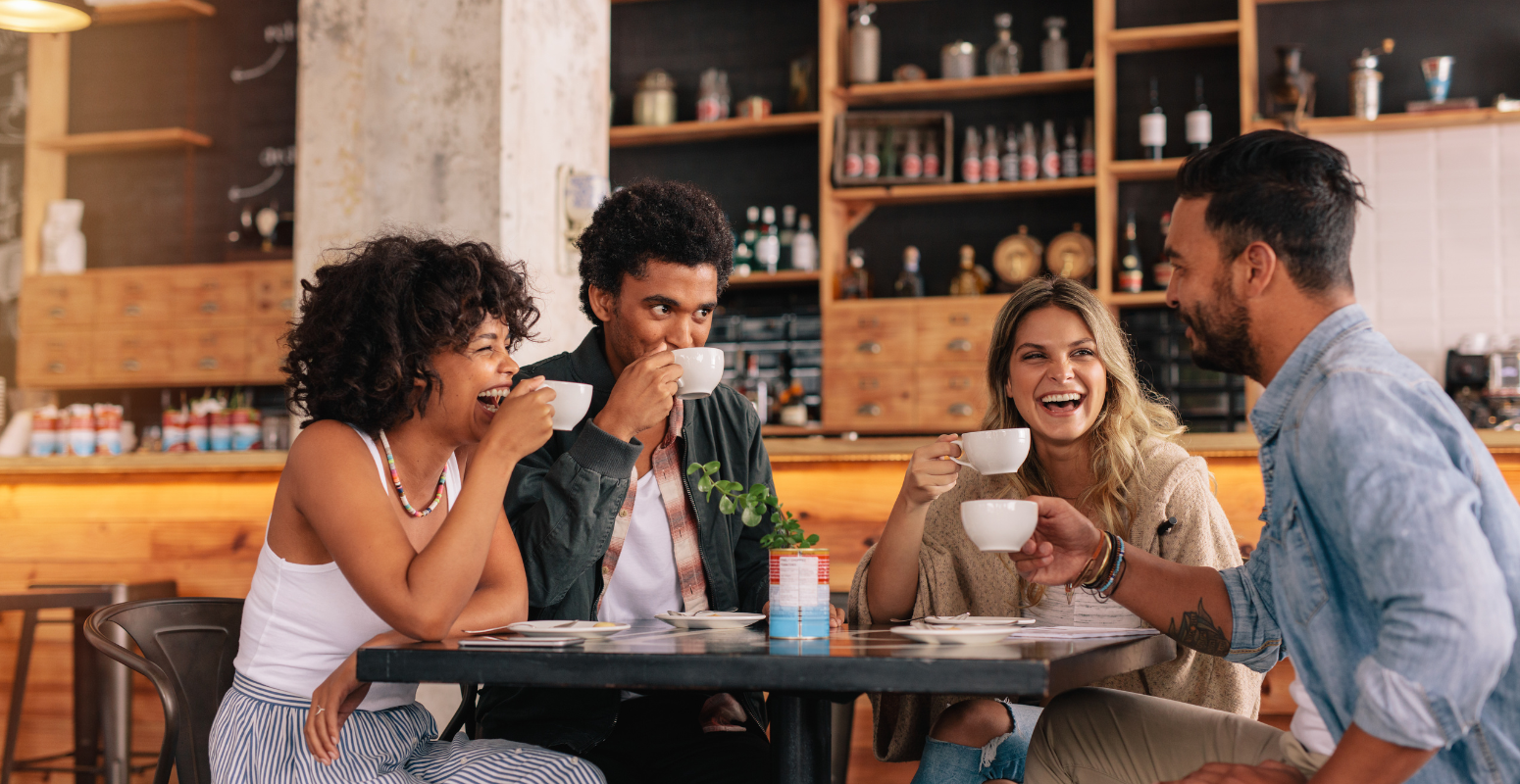 Group of friends laughing and enjoying coffee at a café, symbolizing social connections and engagement in refer-a-friend campaigns.