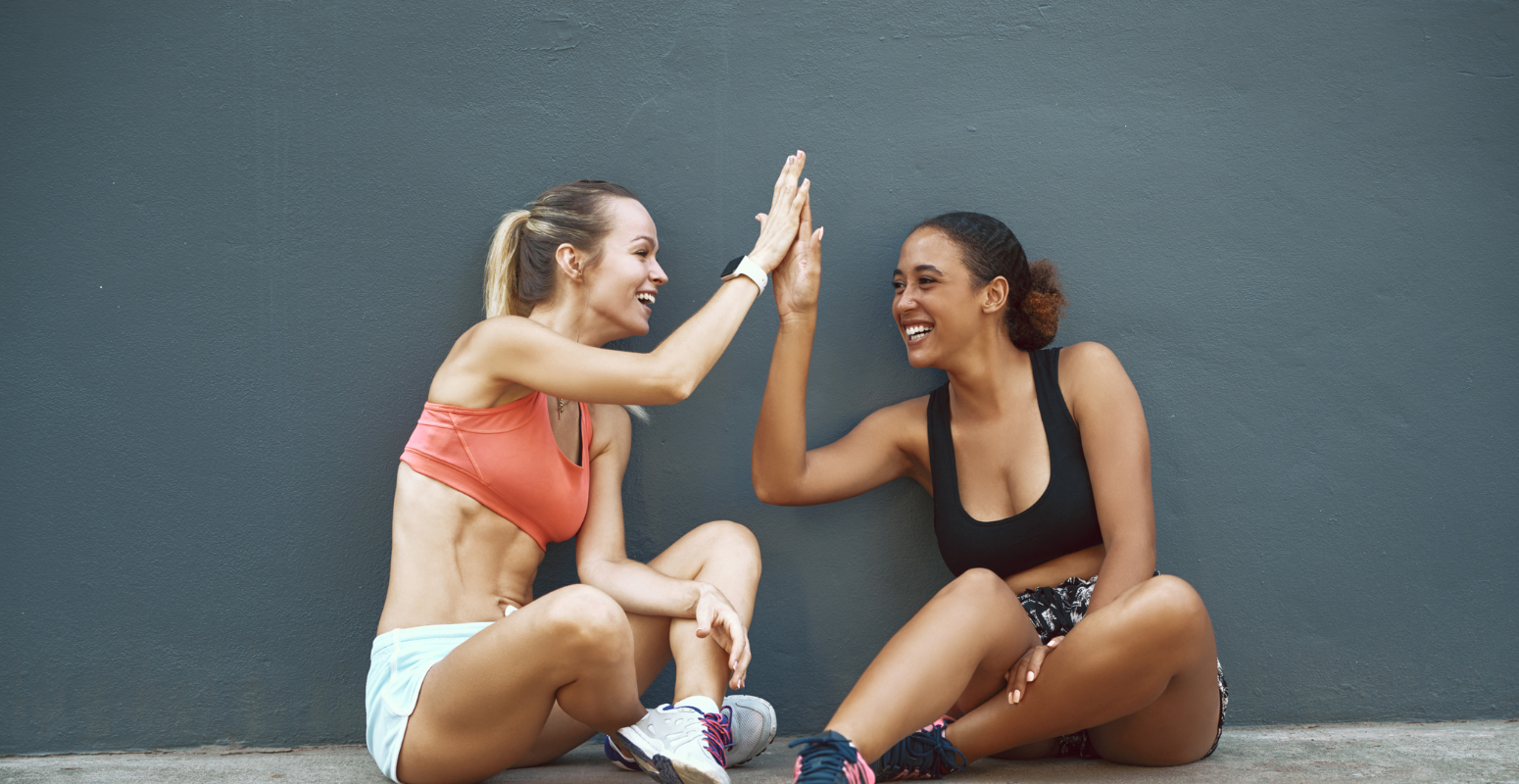 Two fit women in workout gear sitting against a wall, smiling and giving each other a high-five, symbolizing the power of social connections and word-of-mouth recommendations—just like Facebook enhances Referral Marketing through seamless sharing and trust-based engagement.