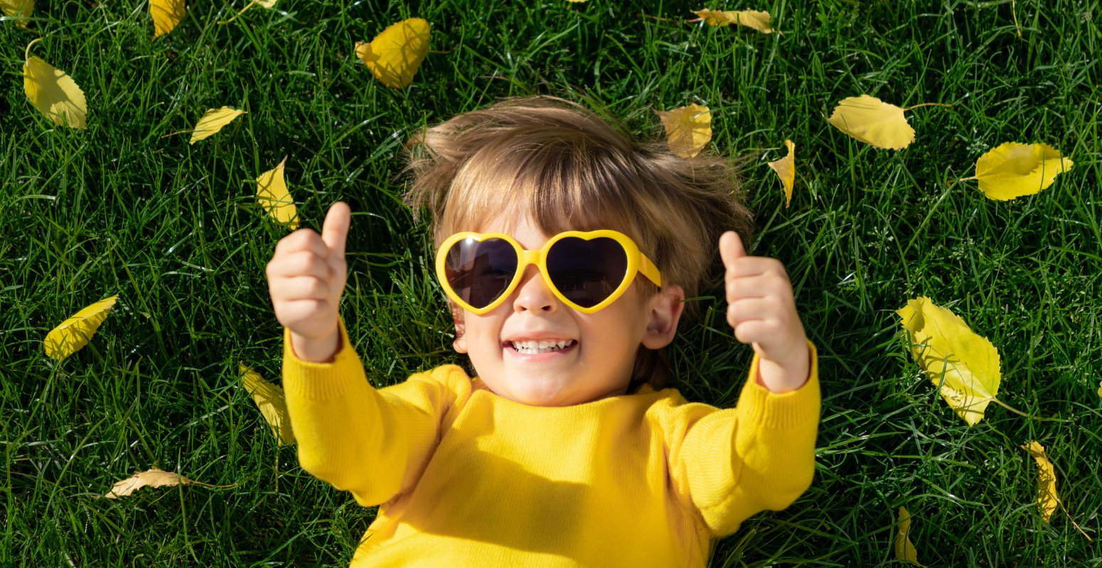 Smiling child in yellow sunglasses and sweater lying on green grass with yellow autumn leaves, giving a thumbs-up.