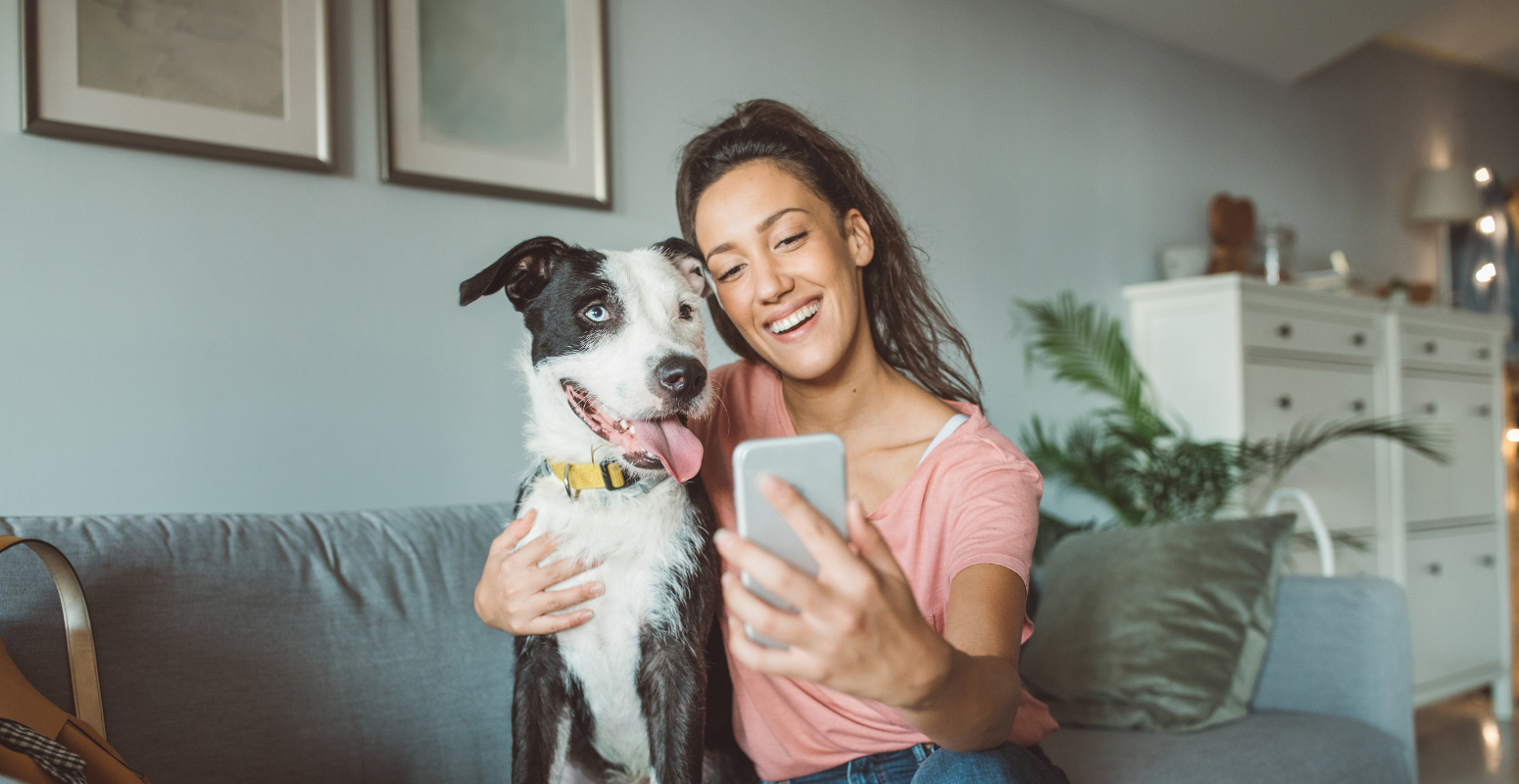 A happy woman sitting on a sofa taking a selfie with her dog, smiling while holding a phone. The cozy living room setting highlights comfort and connection.
